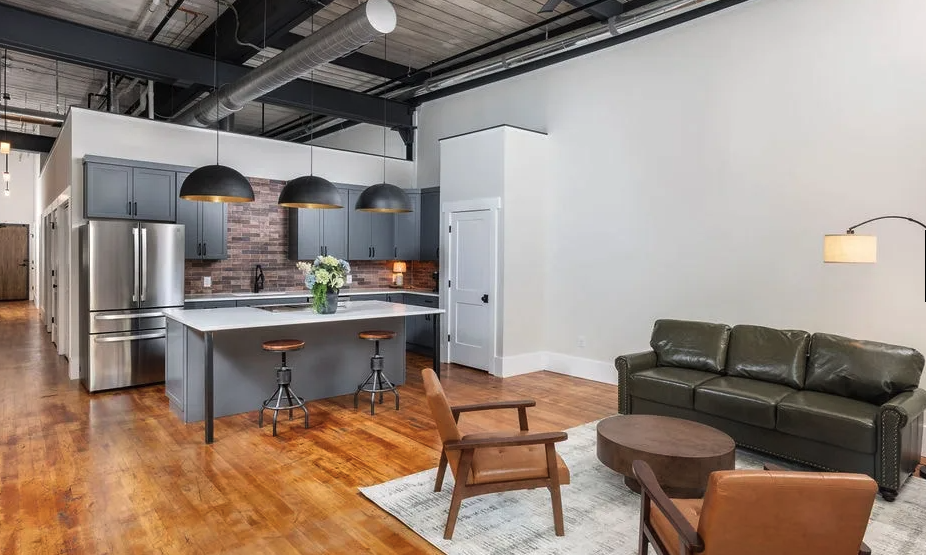Interior of a Lill Mill Lofts apartment in Monroe GA showing open-plan kitchen with quartz island, pendant lighting, brick backsplash, refinished hardwood floors, and living area with leather sofa inside the restored 1900 Walton Cotton Mill
